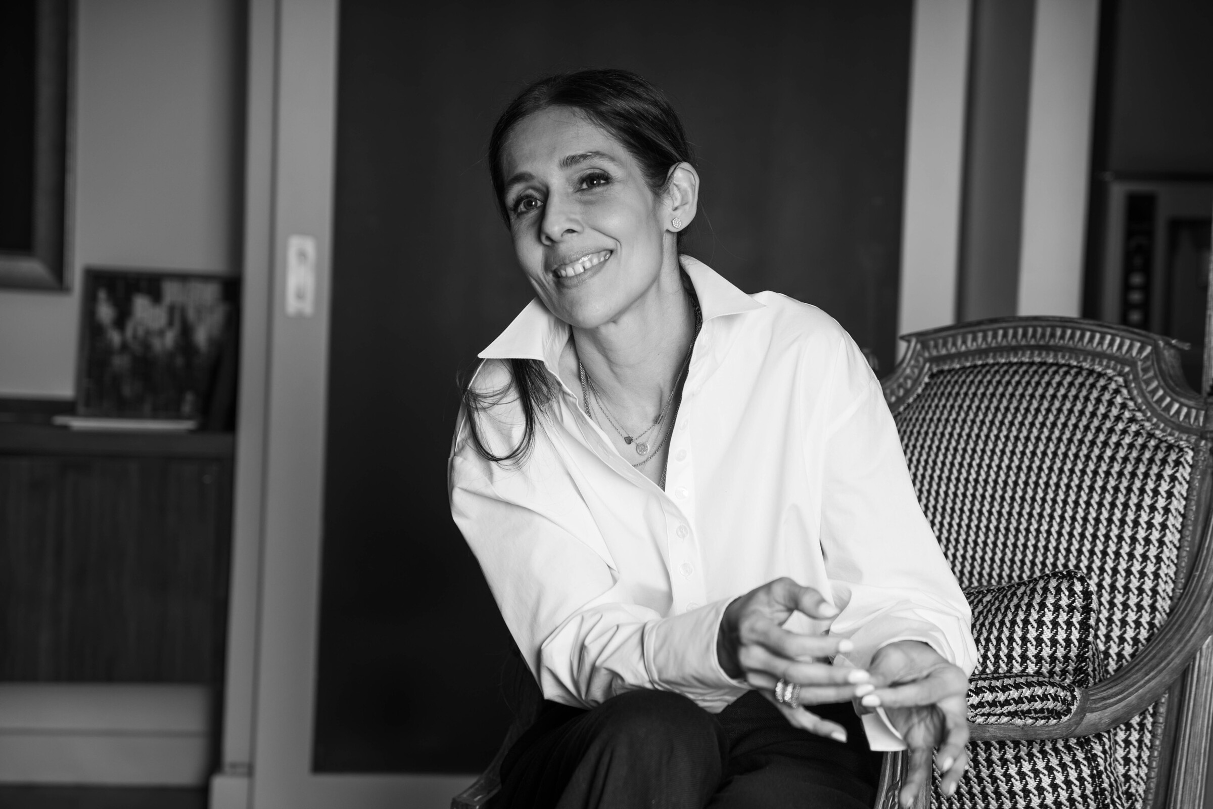 Bushra Khokhar, black and white portrait, seated and smiling in a studio armchair