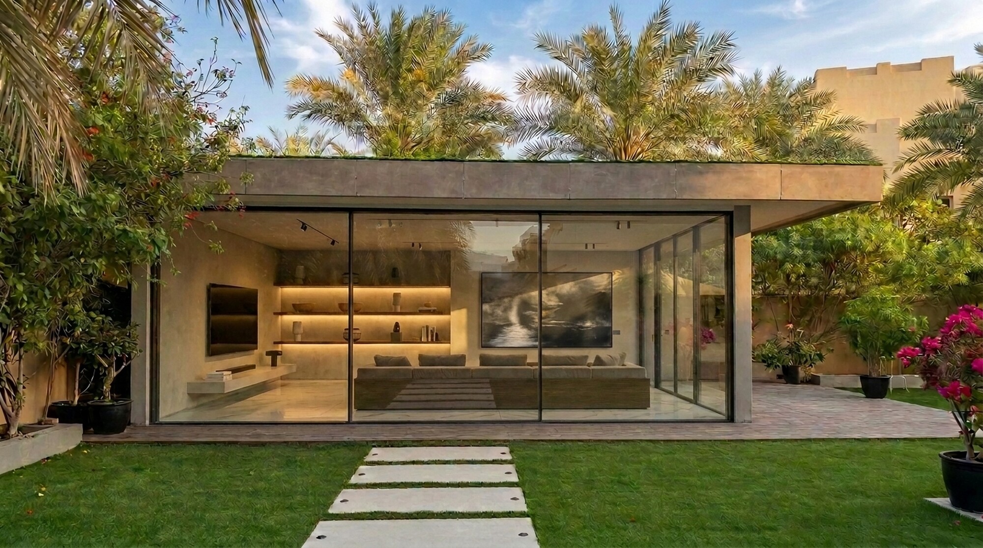 The Hattan annex: a glazed garden pavilion under a deep stone canopy, sliding walls folded open to a lit interior of built-in shelving and a long stone counter, stepping stones across the lawn